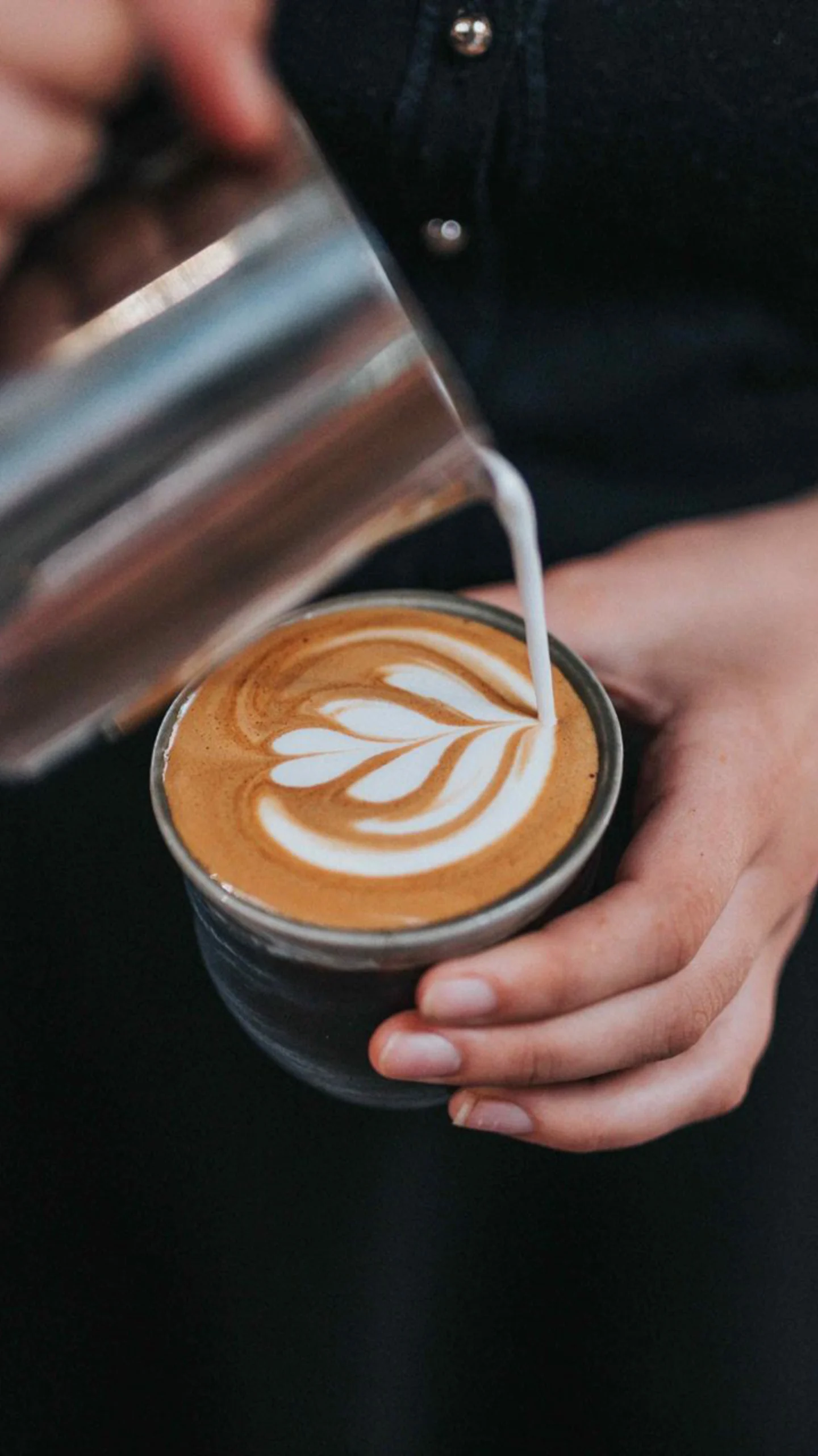 Close-up of a person pouring steamed milk into a cup of coffee to create latte art in the shape of a leaf. The barista is holding a stainless steel milk pitcher and carefully crafting the design in the foam atop the coffee.
